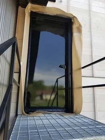 Black door, reflective surface, protected by brown paper, on metal grating, near stairs.