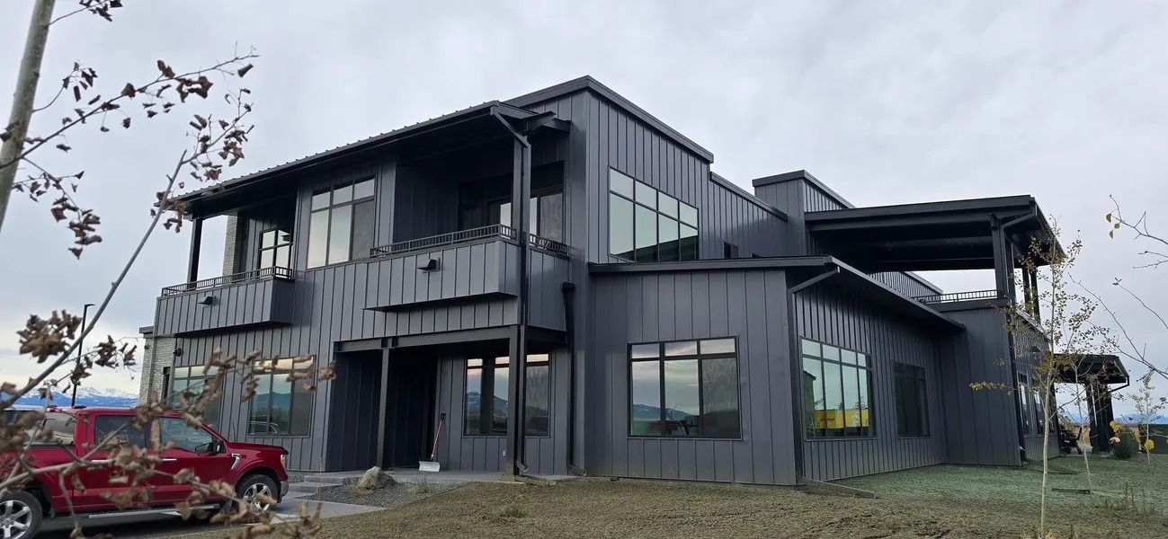 A modern, gray house with large windows and a red truck parked in the yard, set on a cloudy day.