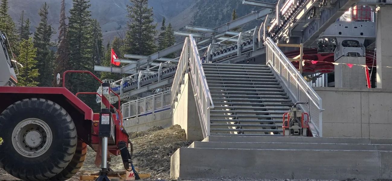 A red tracked vehicle clearing snow from steps leading to a cable car station. Mountain backdrop.