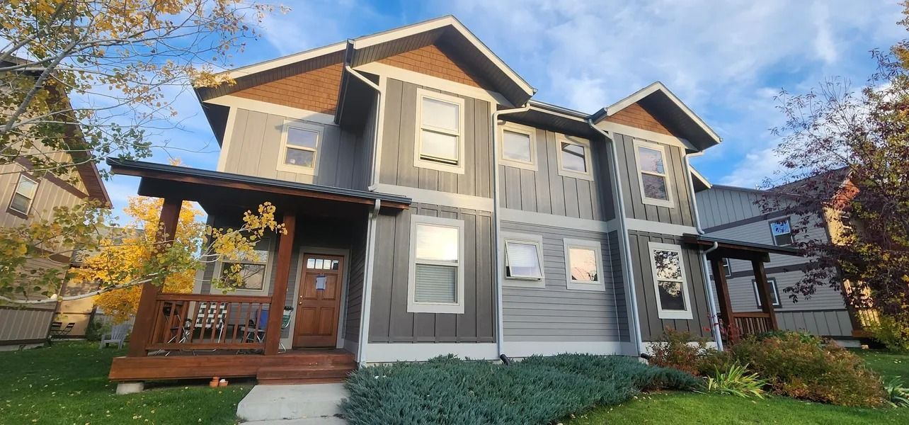 A two-story gray and brown house with a porch and green lawn under a cloudy blue sky.