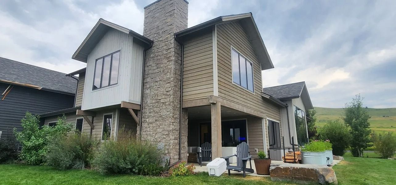 House with stone chimney and siding, surrounded by grass and shrubs, under a cloudy sky.
