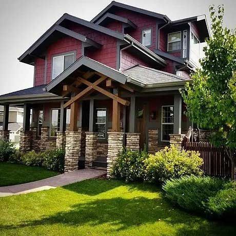 Red and gray two-story house with stone columns, green lawn, and shrubbery. Sunny day.