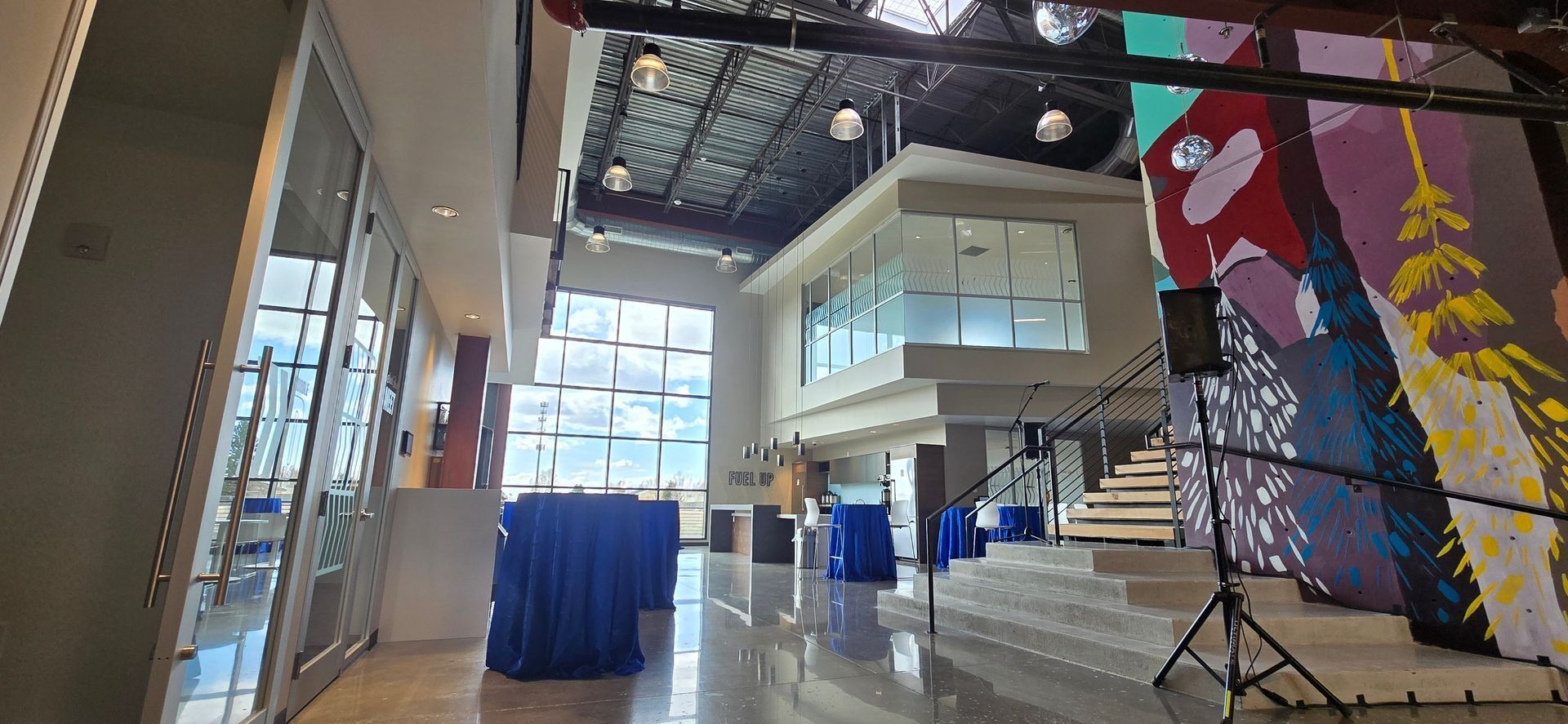 Interior of a modern building with high ceilings, glass windows, and a staircase. Tables covered in blue cloth and a colorful mural are visible.