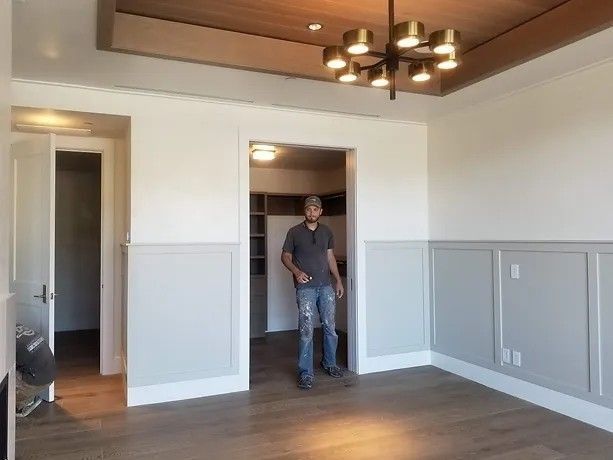 Man standing in a room with wood floors, molding, and a chandelier. Doorways lead to other rooms.