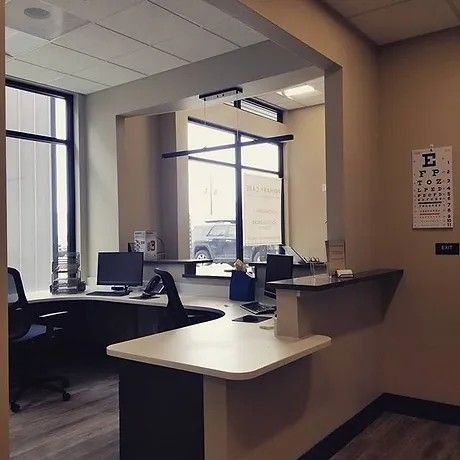 Reception area of a medical office with a desk, computers, and an eye chart. Window looks outside.