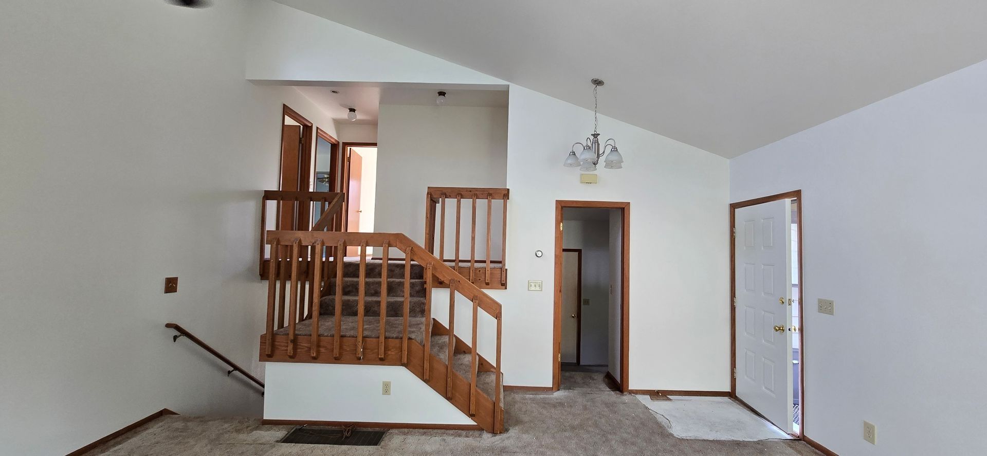 Interior view of a home's entryway. Wooden staircase and railing, doorway to the left, and a chandelier in the center.