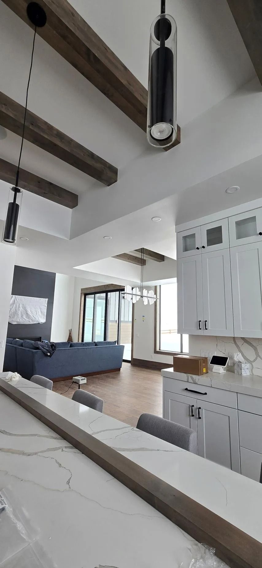 Kitchen interior with white cabinets, dark wood beams, and pendant lights. A view of a living area is visible.
