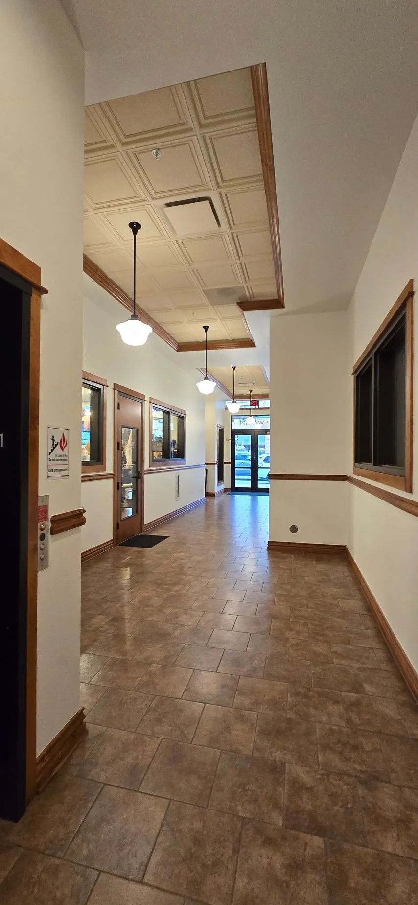 Hallway with tile floor, cream walls, and overhead lights. Doors and windows line both sides.