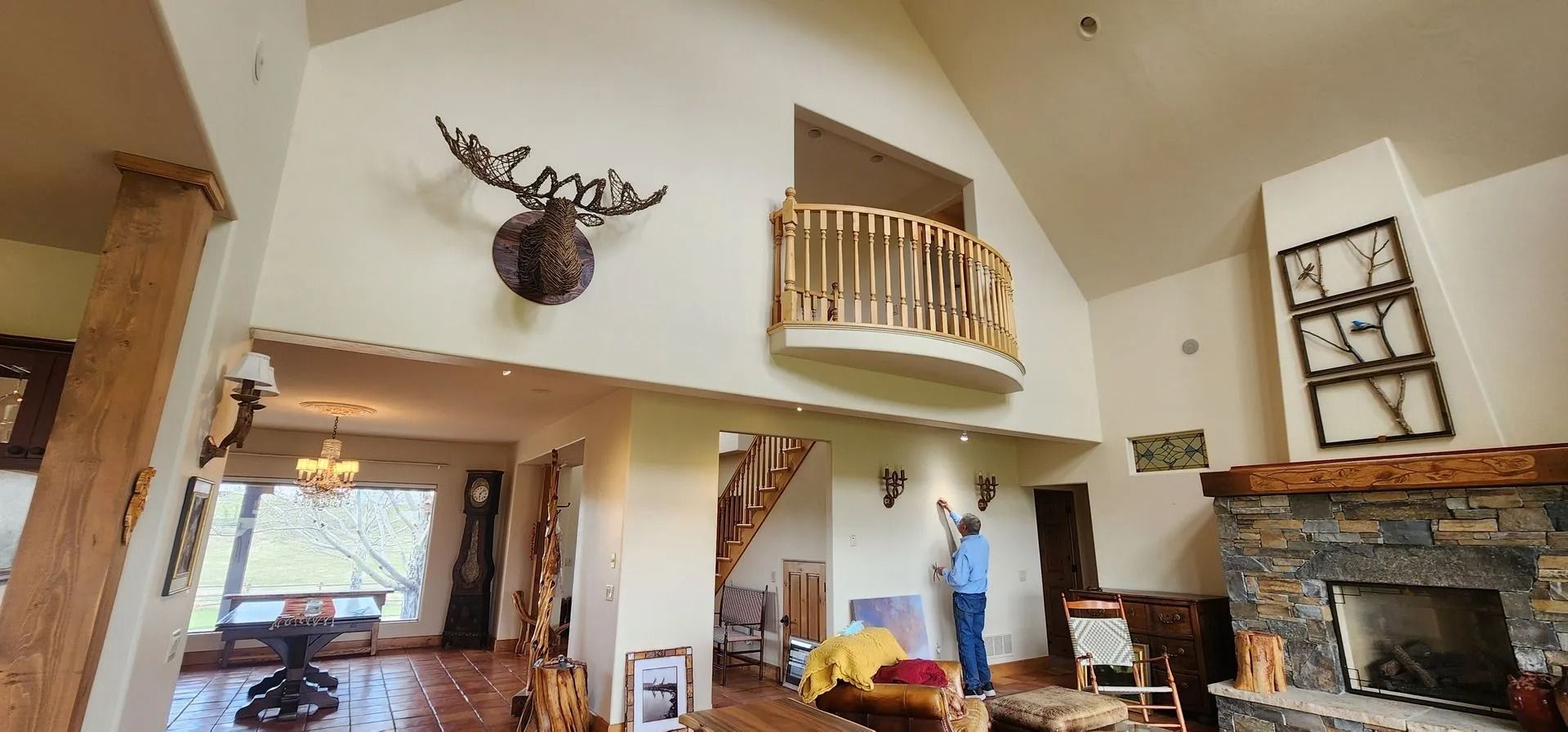 Spacious room with high ceilings, fireplace, balcony, and mounted elk head. Person painting on wall.