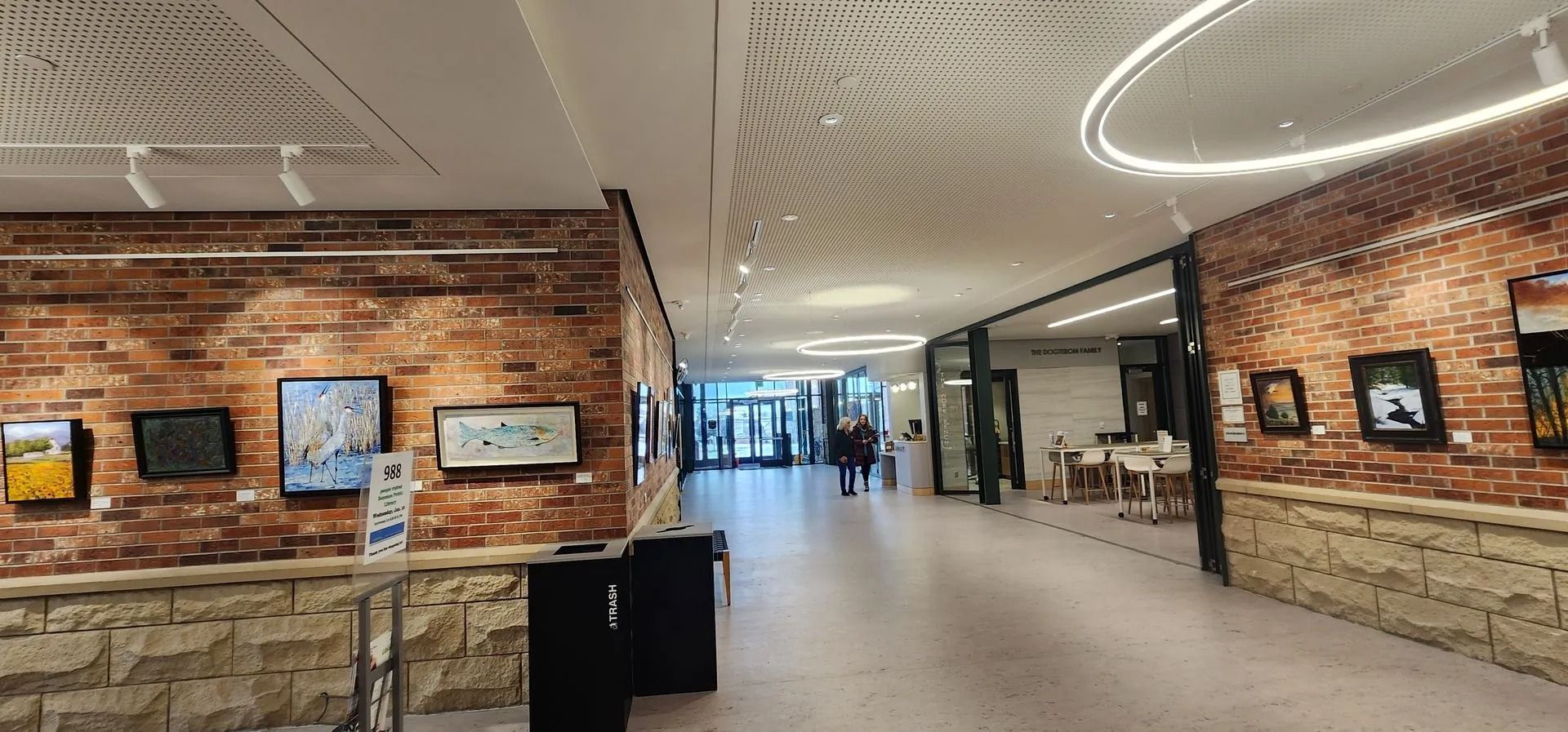 Hallway with brick walls, art, and people walking toward a glass entrance.