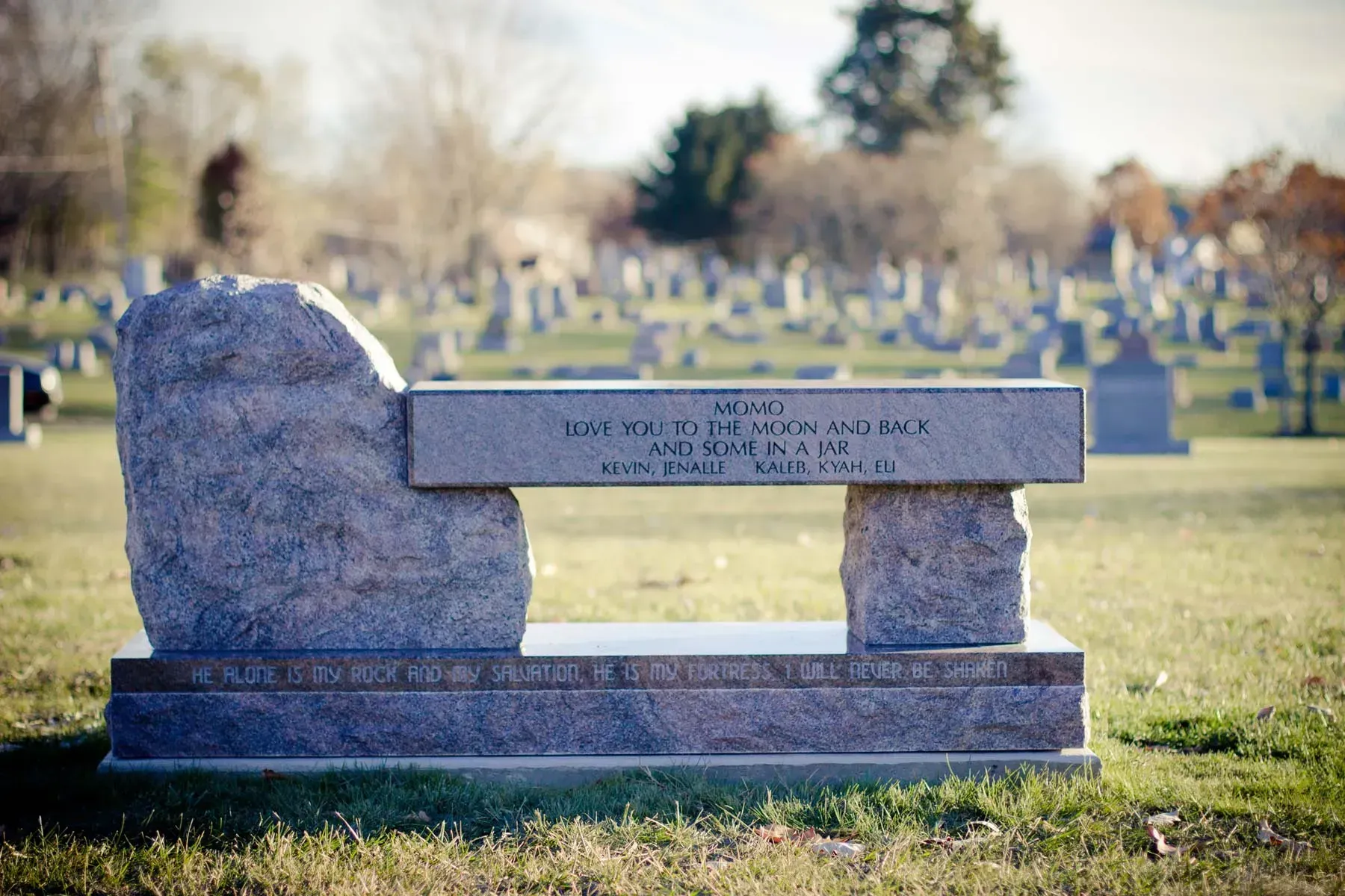 Stone bench-style memorial marker in a cemetery, with gravestones blurred in the background.