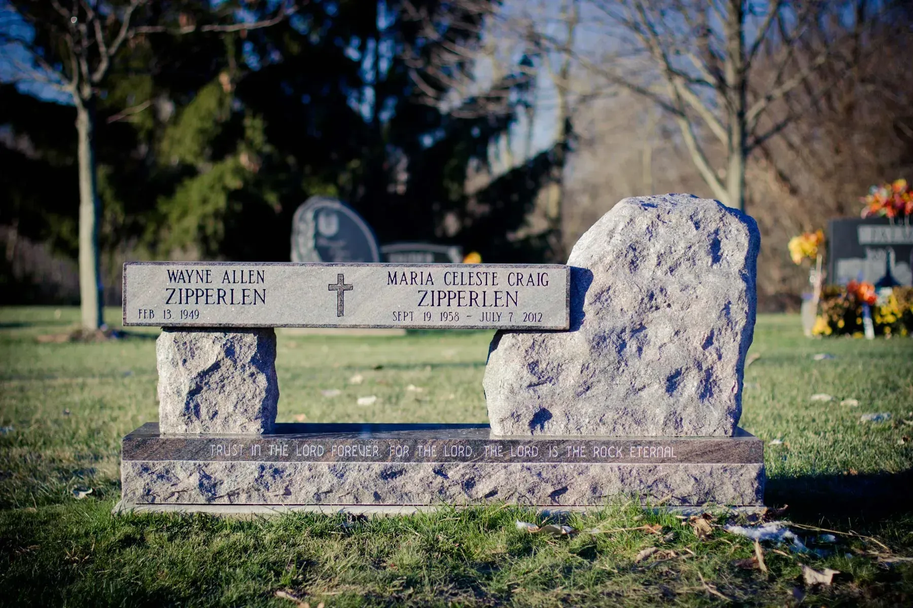 Granite memorial bench in a cemetery with engraved names and a floral arrangement under it.