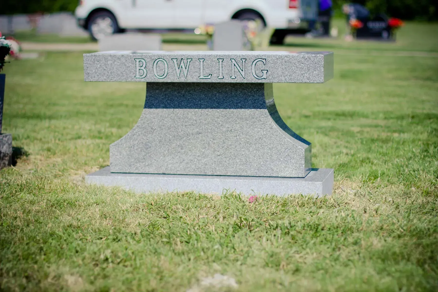 Gray gravestone engraved “BOWLING” in a grassy cemetery, with a blurred car in the background