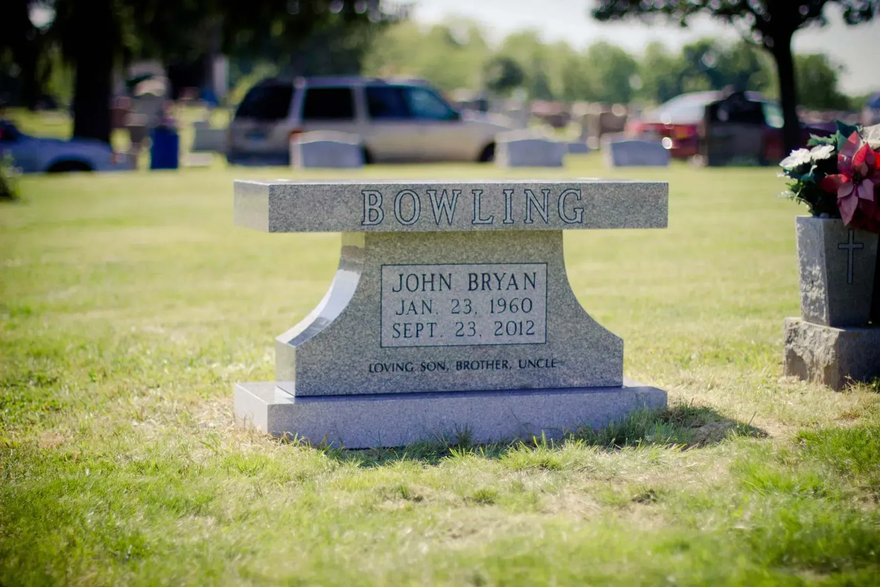 Gravestone in a grassy cemetery, with flowers nearby and blurred vehicles in the background.