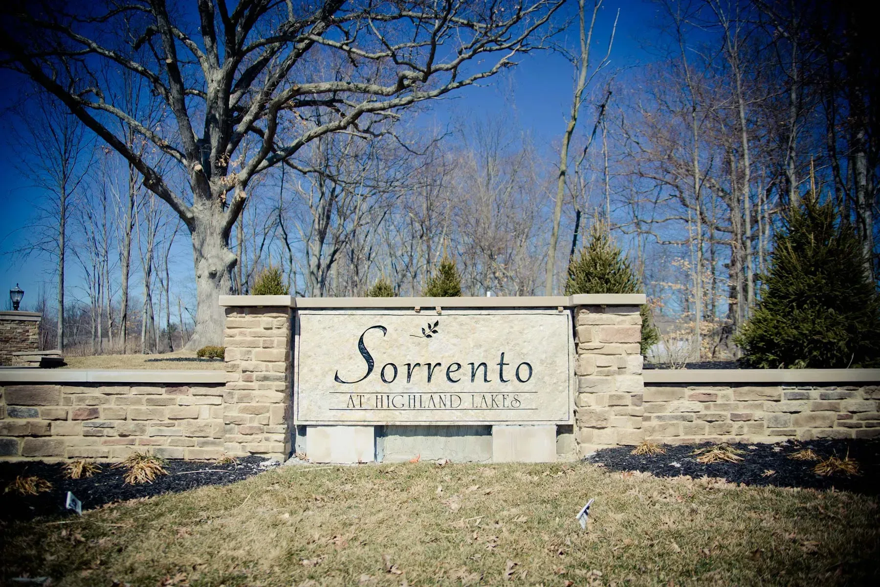 Stone entrance sign reading “Sorrento” in a landscaped outdoor area with trees and blue sky