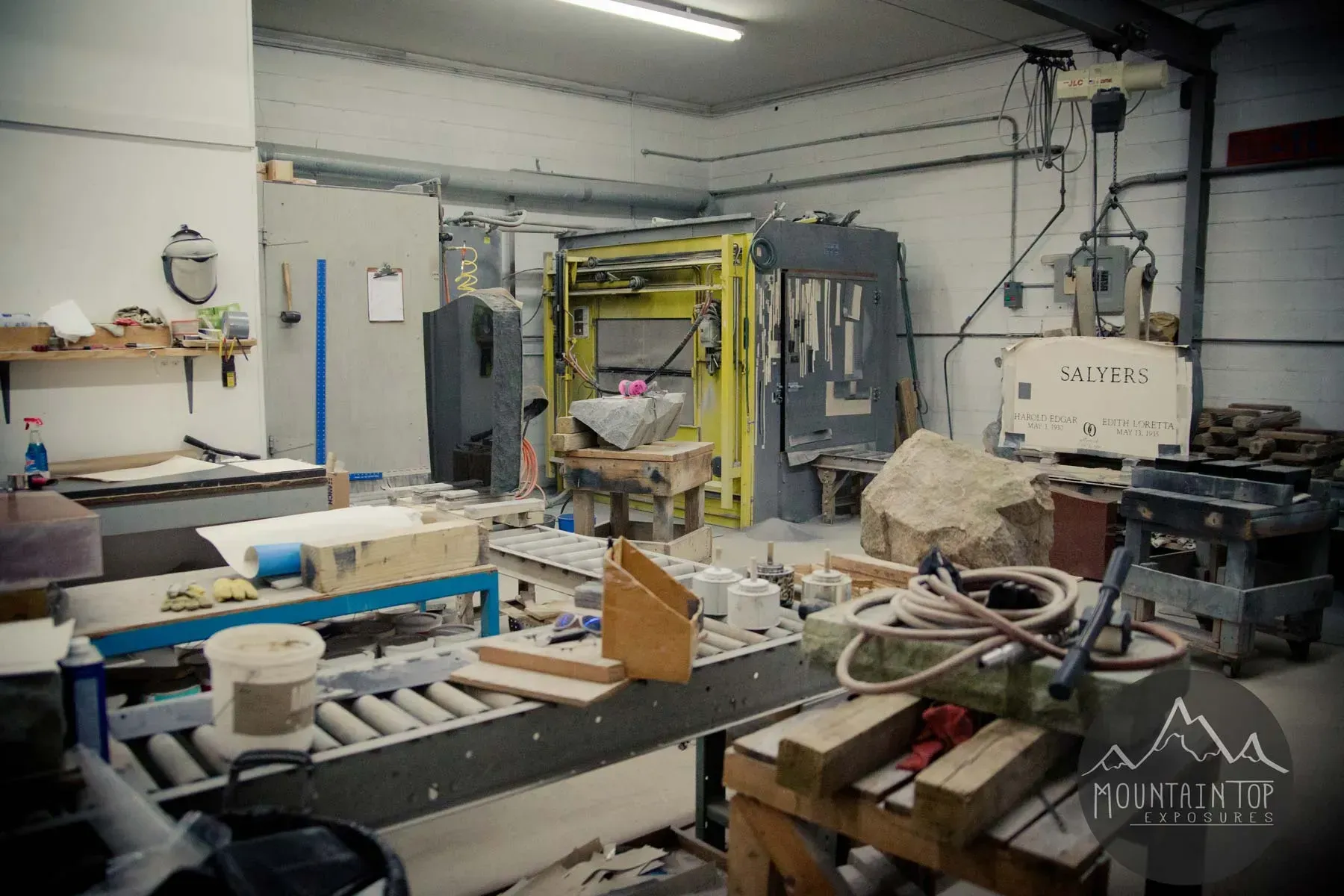 Cluttered workshop with machinery, tables, tools, and materials under bright fluorescent lights