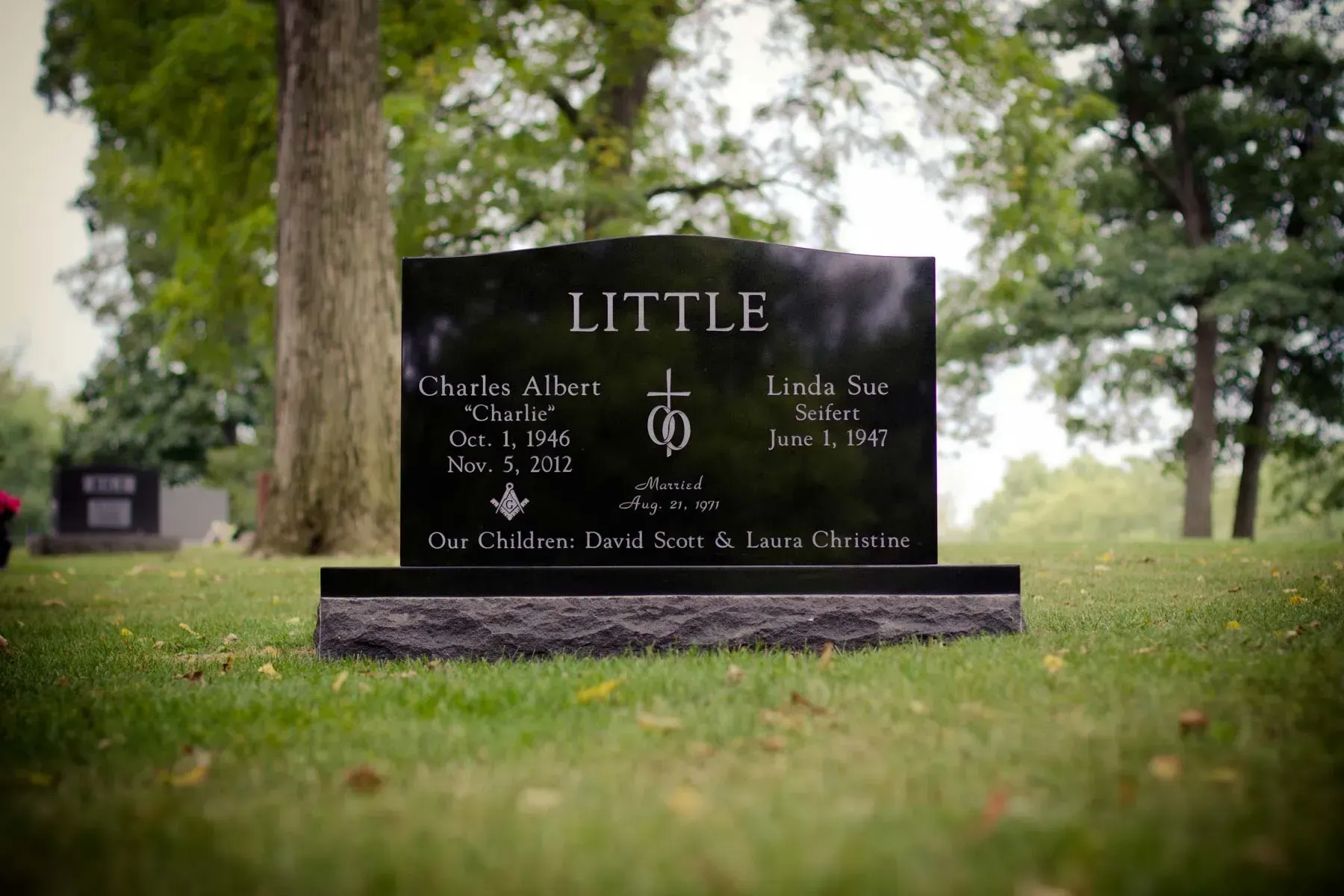 Black memorial gravestone in a grassy cemetery with trees in the background.