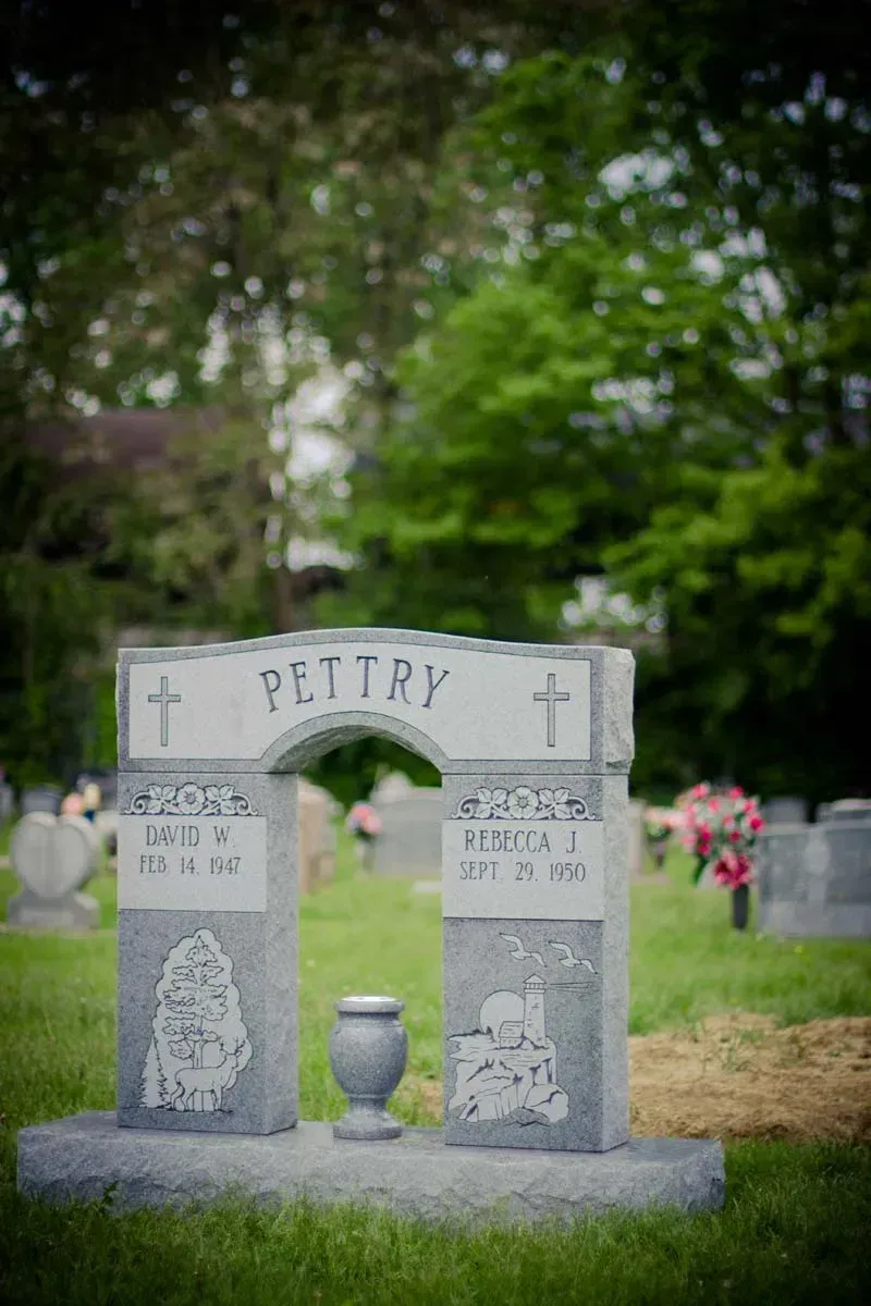 Gray gravestone memorial with “PESTRY” engraved, standing in a cemetery with trees and flowers.