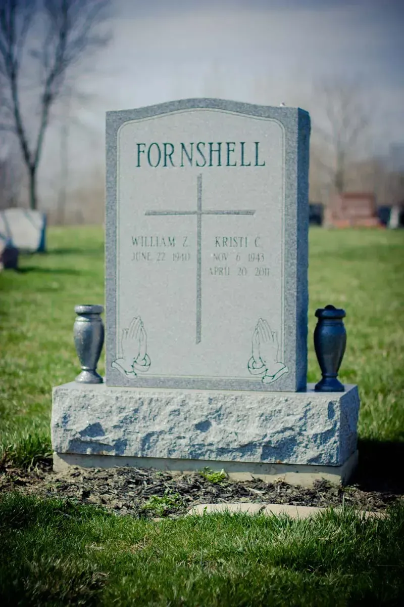 Fornshell gravestone in a cemetery, with gray stone and dark decorative vases on either side.