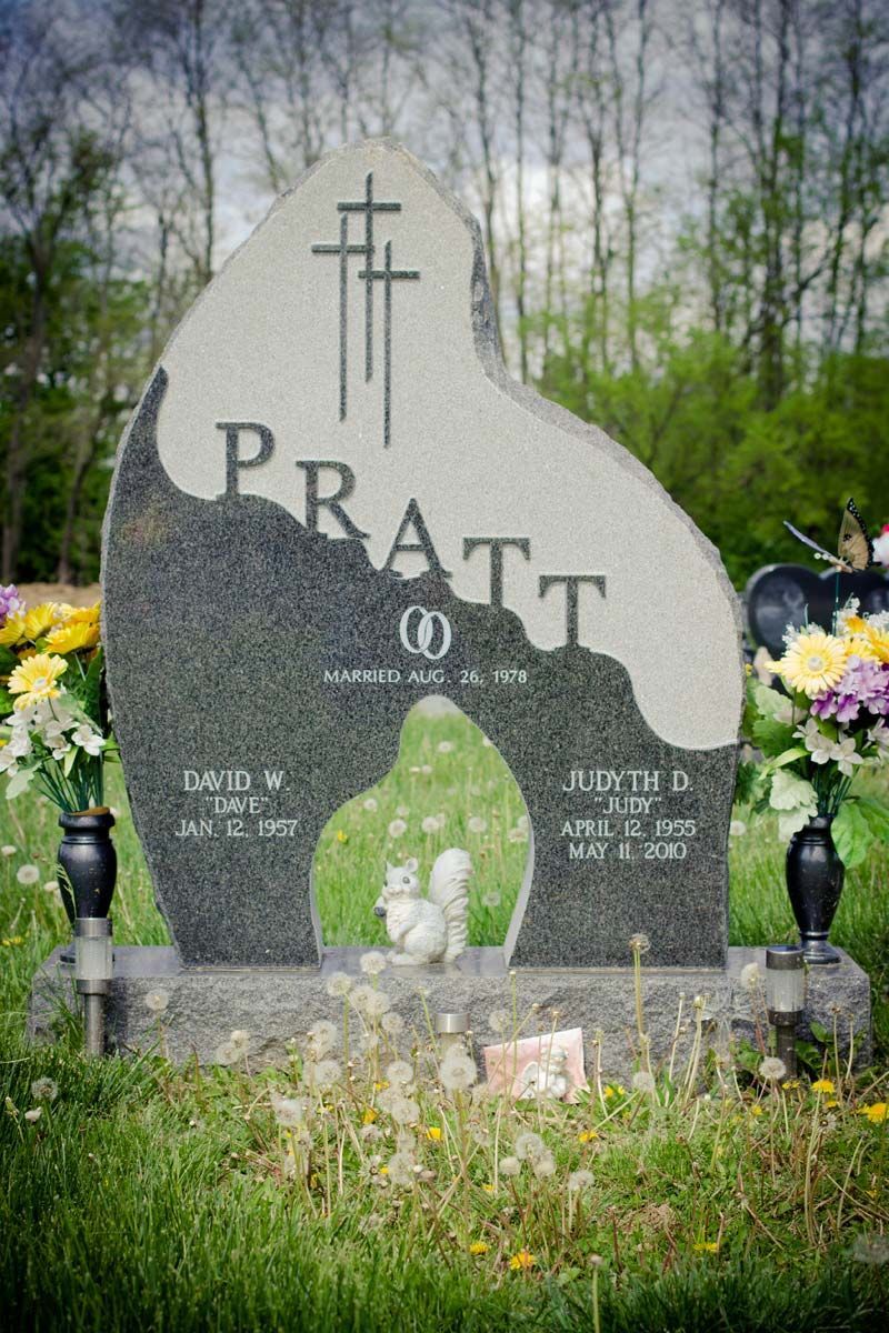 Gray gravestone with cross and PRATT engraved, surrounded by flowers in a cemetery setting