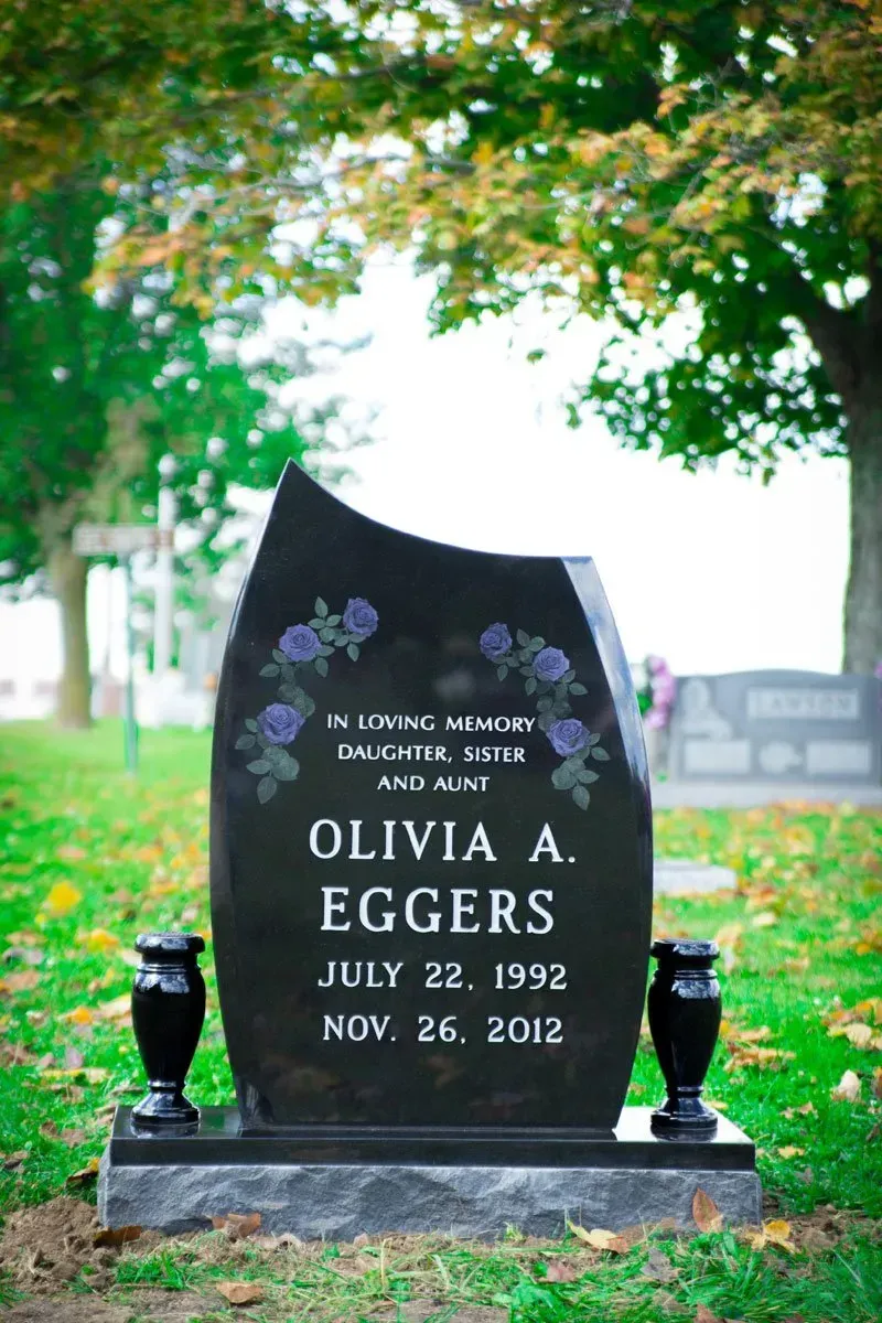 Black gravestone with flowers and vase in a cemetery under trees, inscribed “Olivia A. Eggers” with dates.