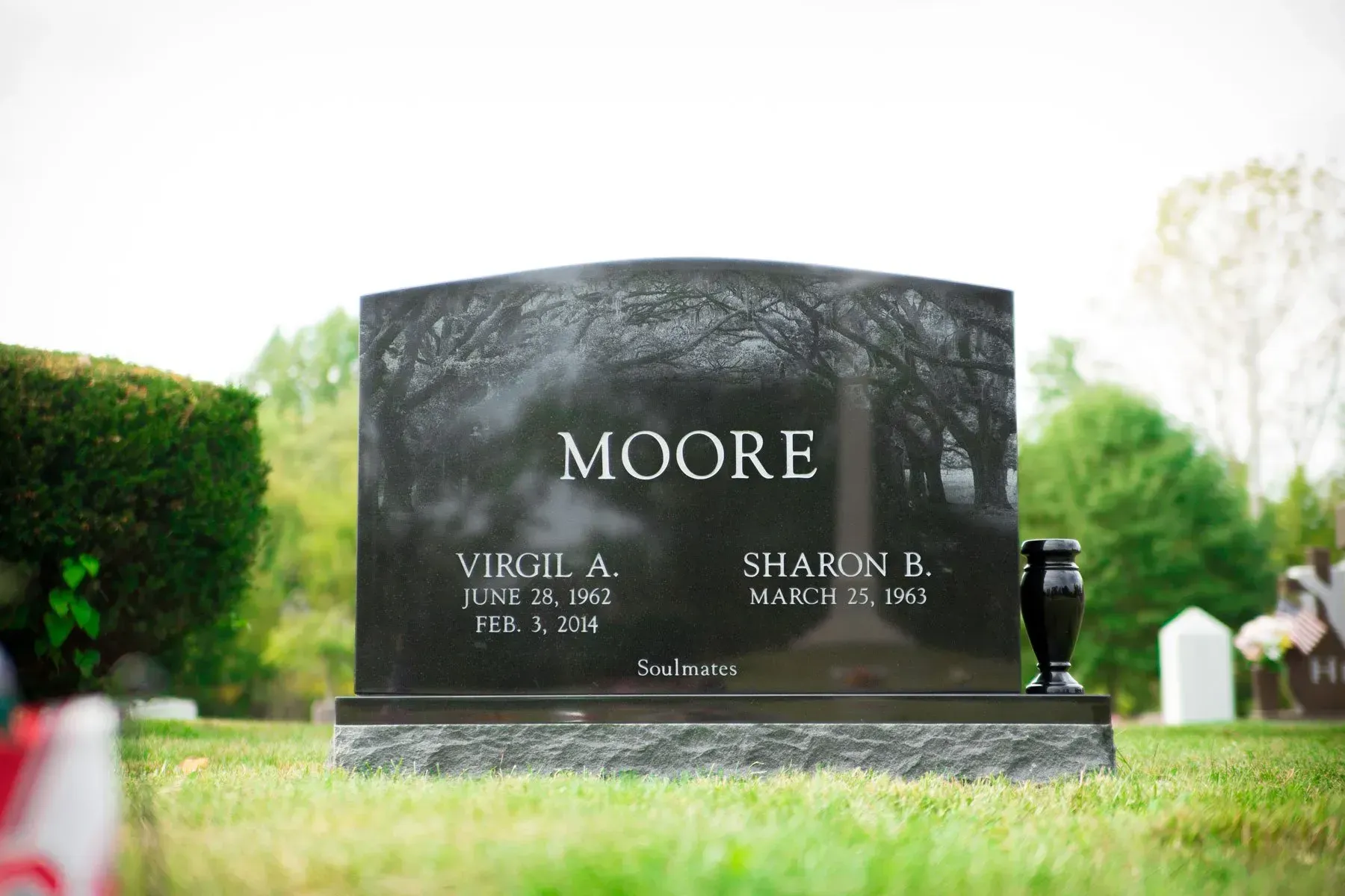 Black granite Moore gravestone in a grassy cemetery with blurred greenery in the background