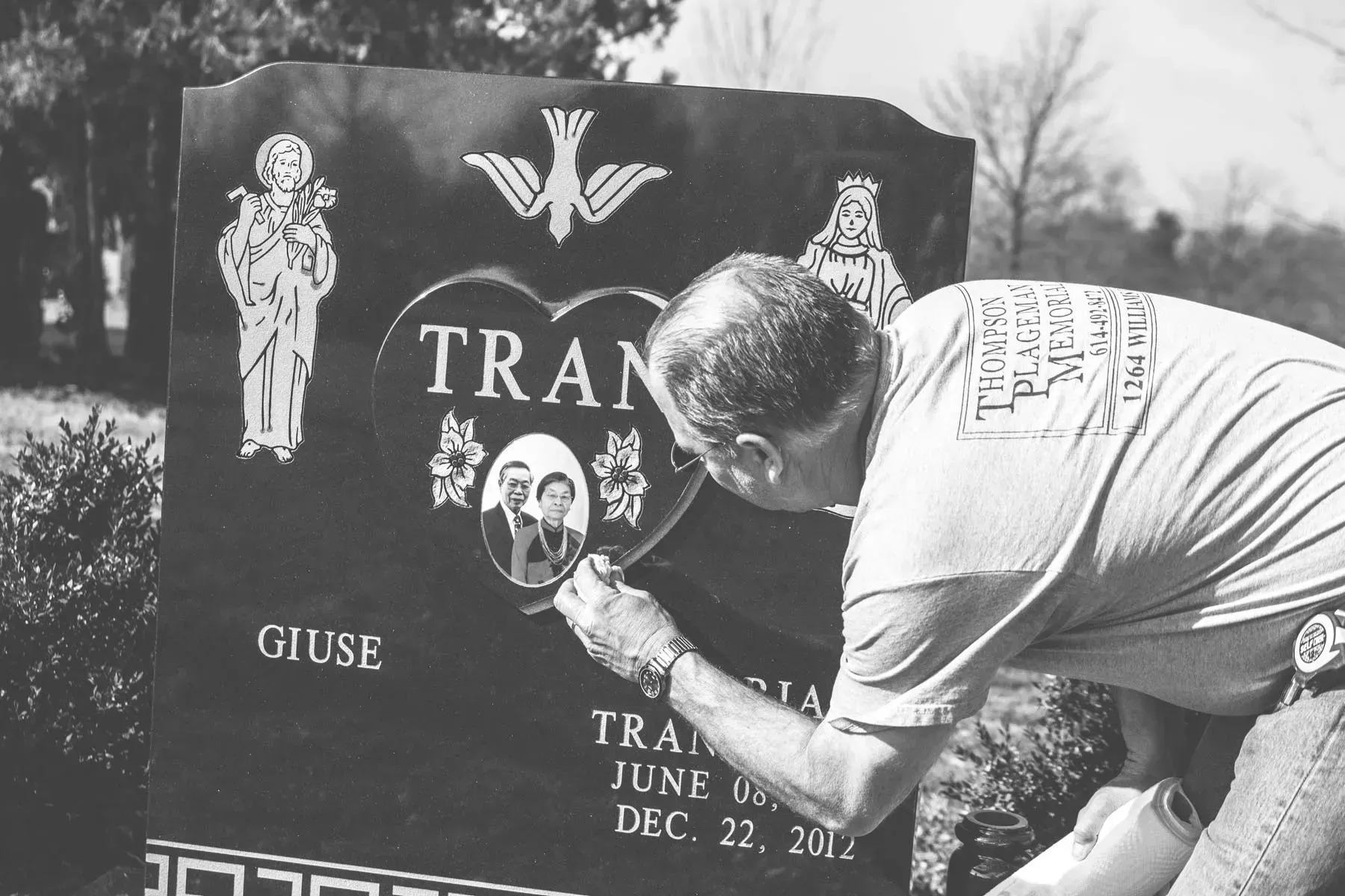 Man kneels at a memorial stone, arranging flowers beneath engraved names and symbols.