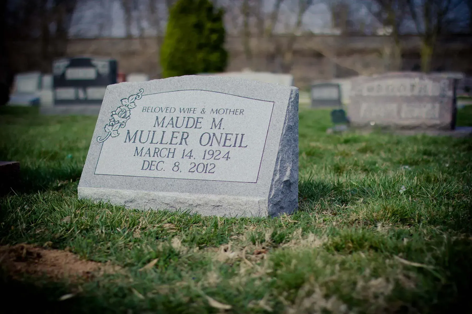 Gravestone in a grassy cemetery, with blurred headstones in the background.
