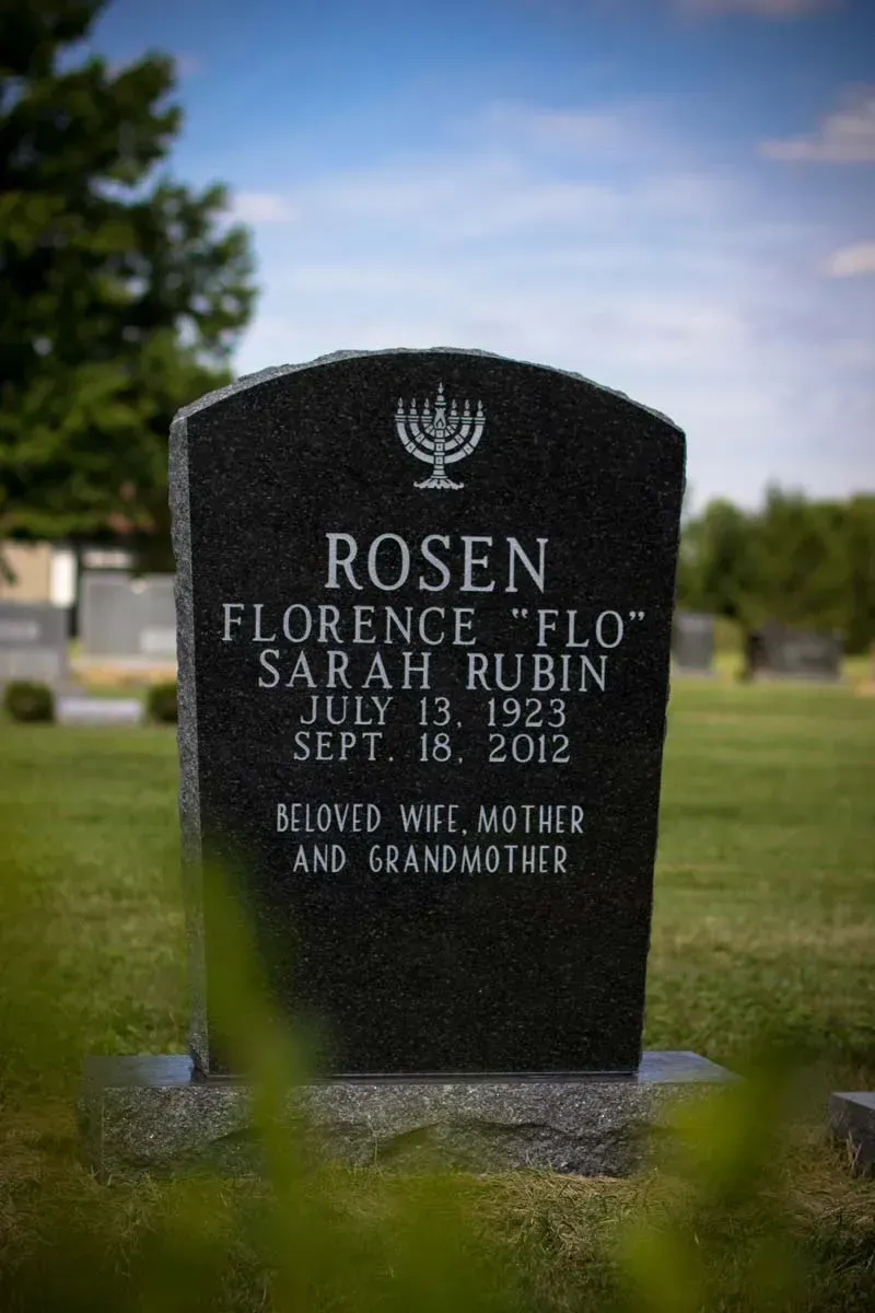 Black gravestone with Hebrew symbol, engraved “ROSEN,” in a cemetery with blurred trees and blue sky.