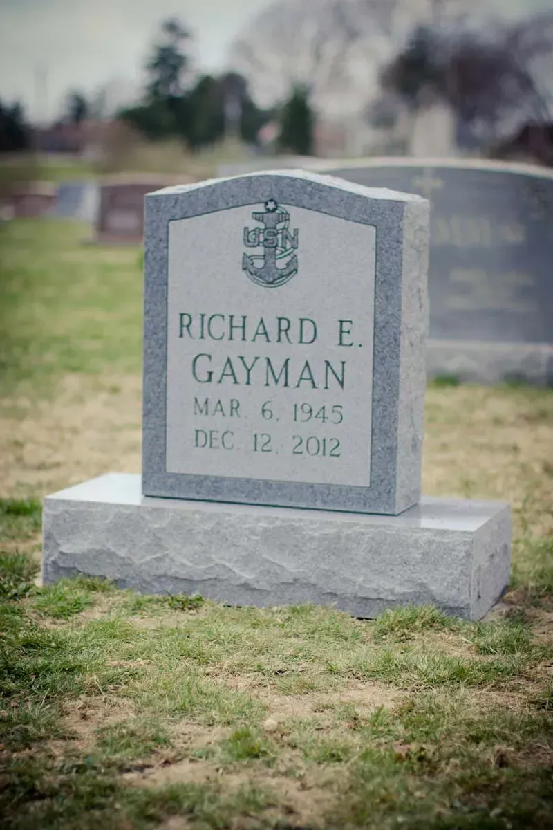 Gray gravestone with inscription “Richard E. Gayman,” standing in a cemetery lawn.