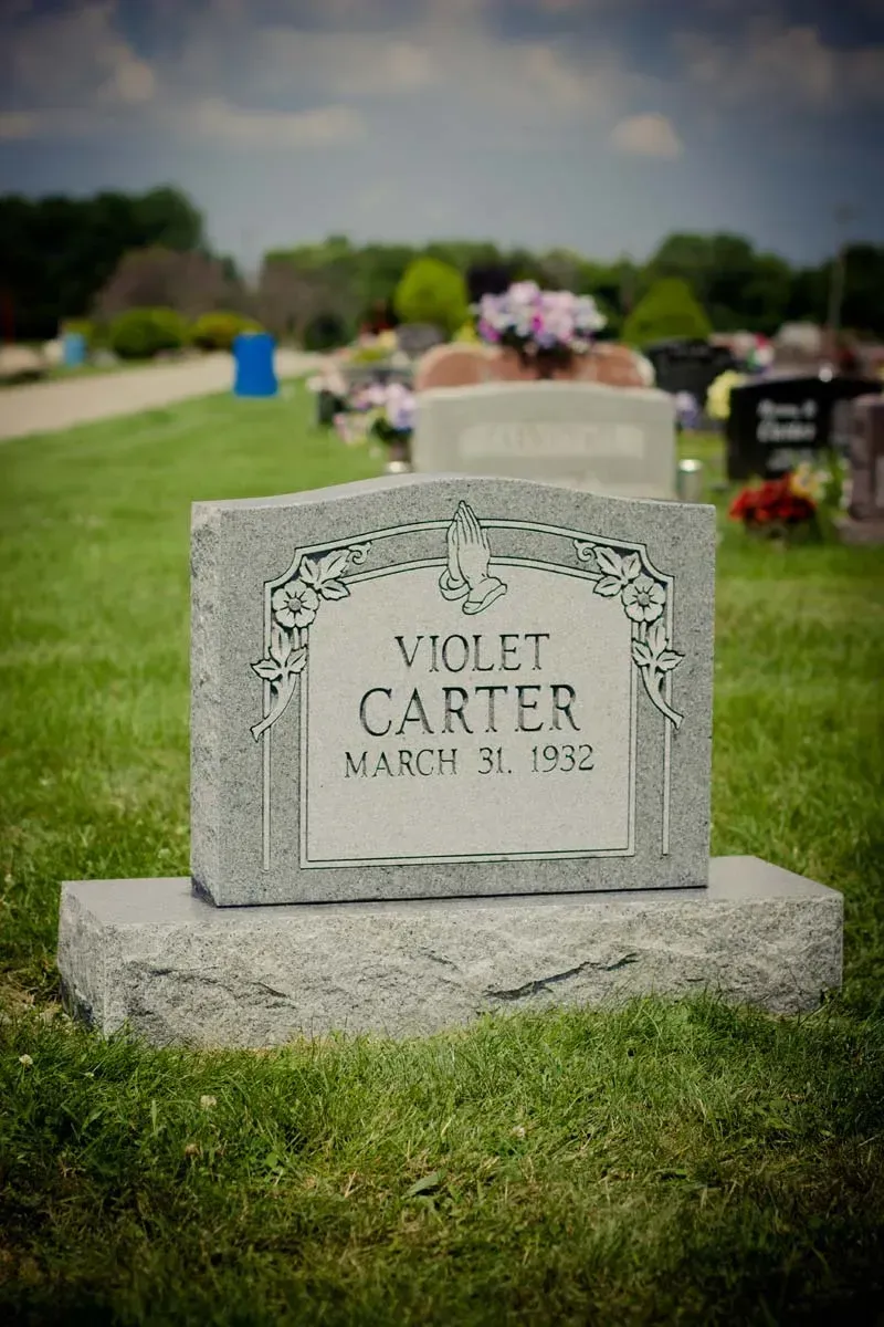 Gray gravestone engraved “Violet Carter” in a cemetery, with grass and blurred flowers behind it