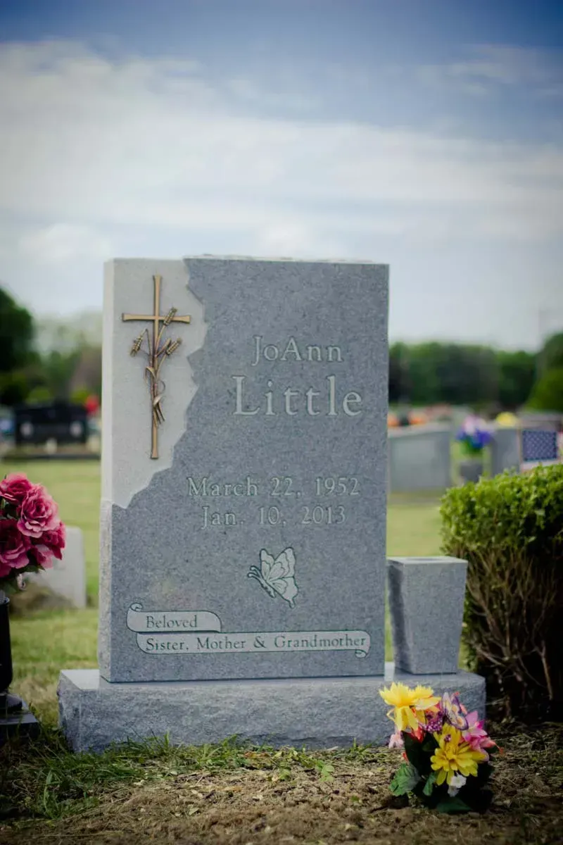 Gravestone with a cross in a cemetery, surrounded by flowers under a cloudy sky