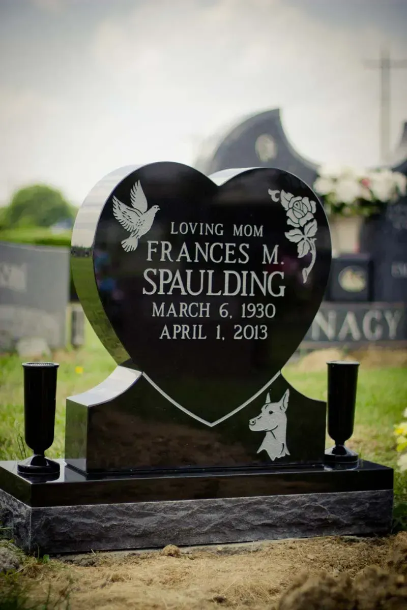 Heart-shaped black gravestone with white inscription in a cemetery, flanked by two small vases.