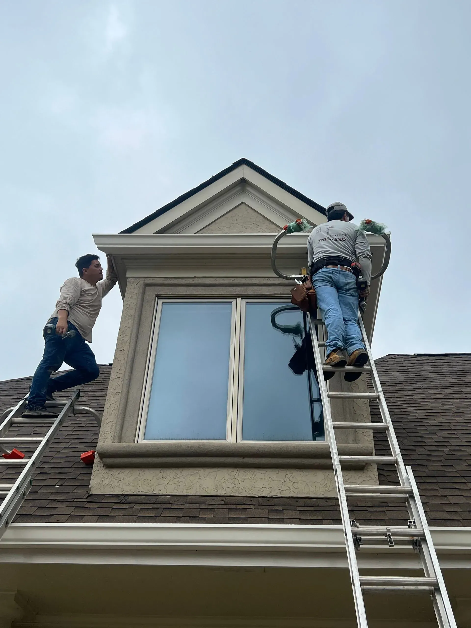 Two workers on ladders repair a roof window under a cloudy sky.