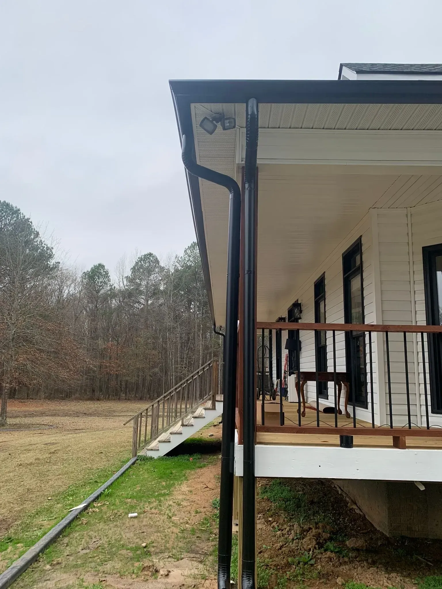 Brown gutters on a white house with a porch and black railing, overlooking a grassy yard and trees.