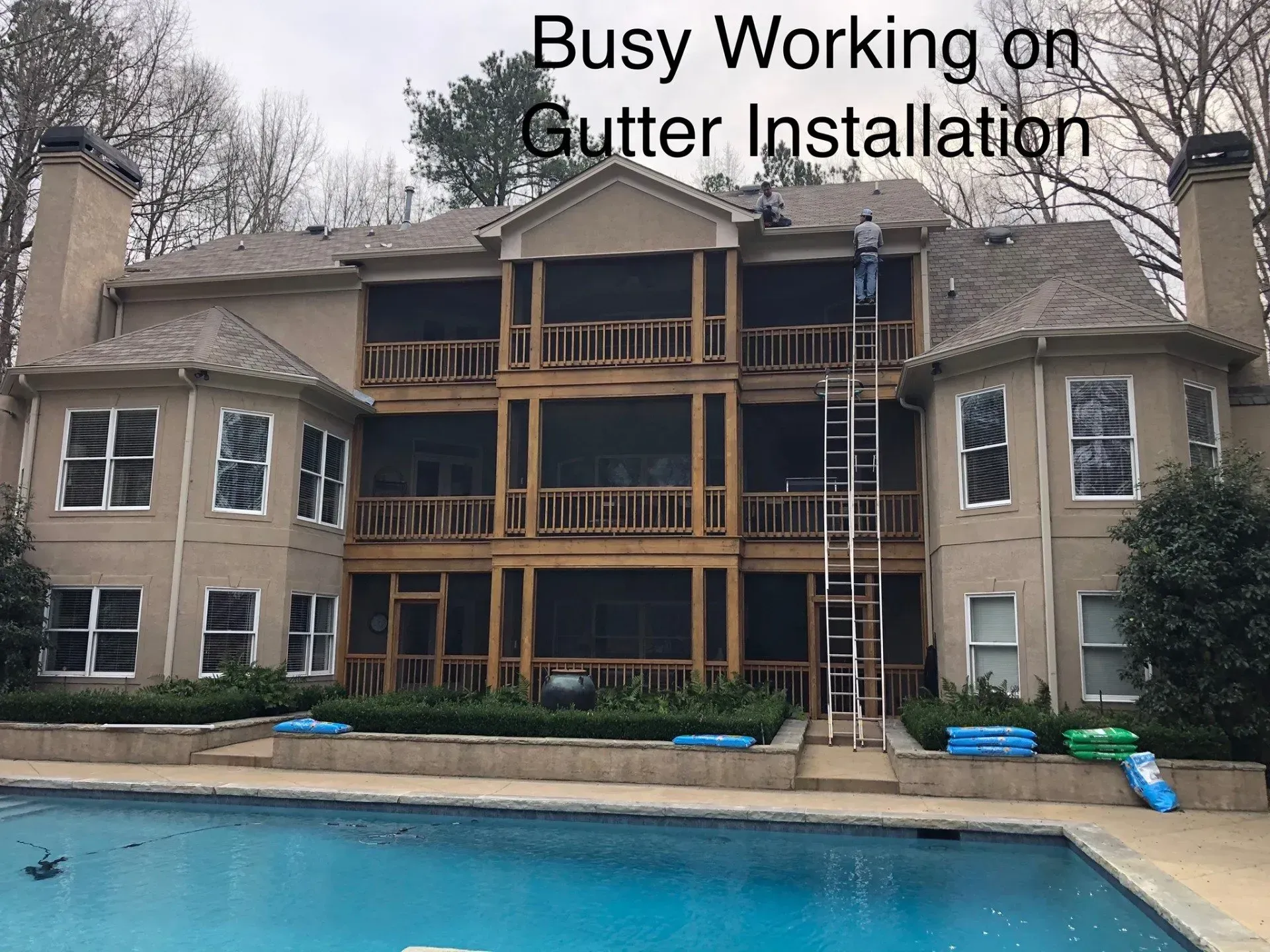 Man on ladder installing gutters on a large house with screened porches near a pool.