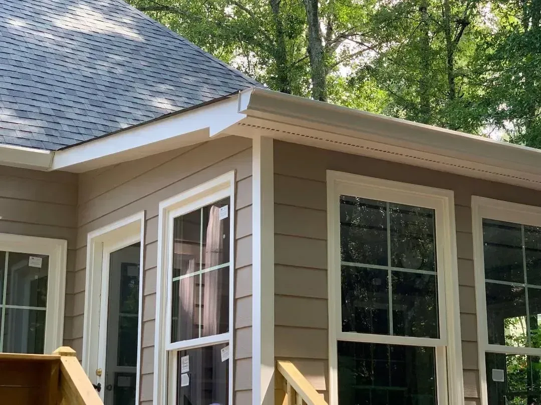 Beige house exterior with windows, roof, and trees in background.
