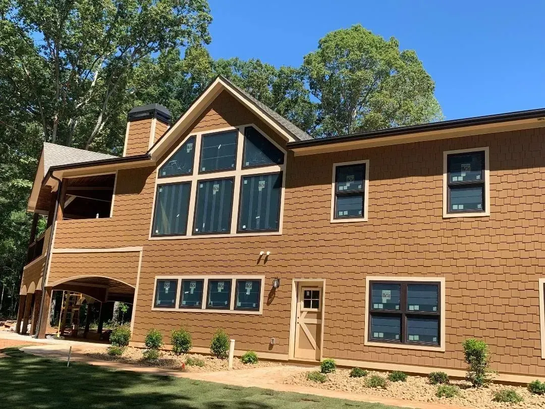 Two-story house with brown textured siding, large windows, and a covered porch, under construction in a wooded area.