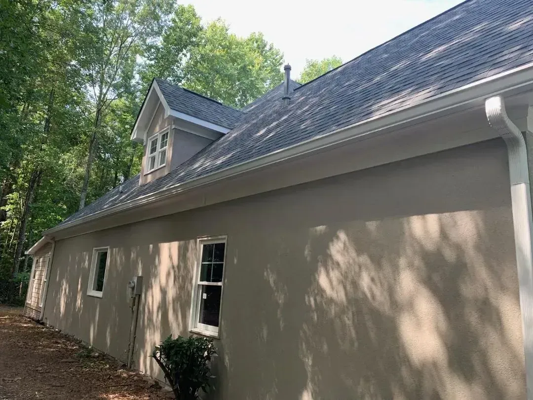 Beige house exterior with gray roof, white gutters and trim, and trees in background.