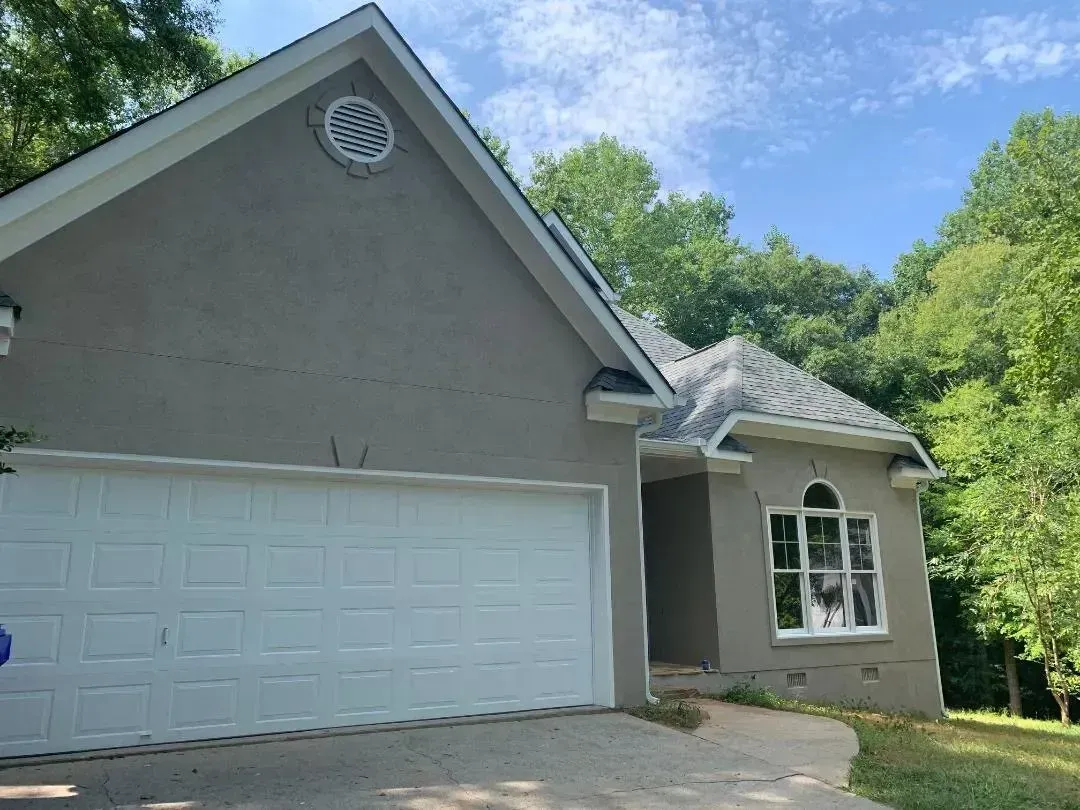 Gray stucco house with white garage door and roof, surrounded by green trees.
