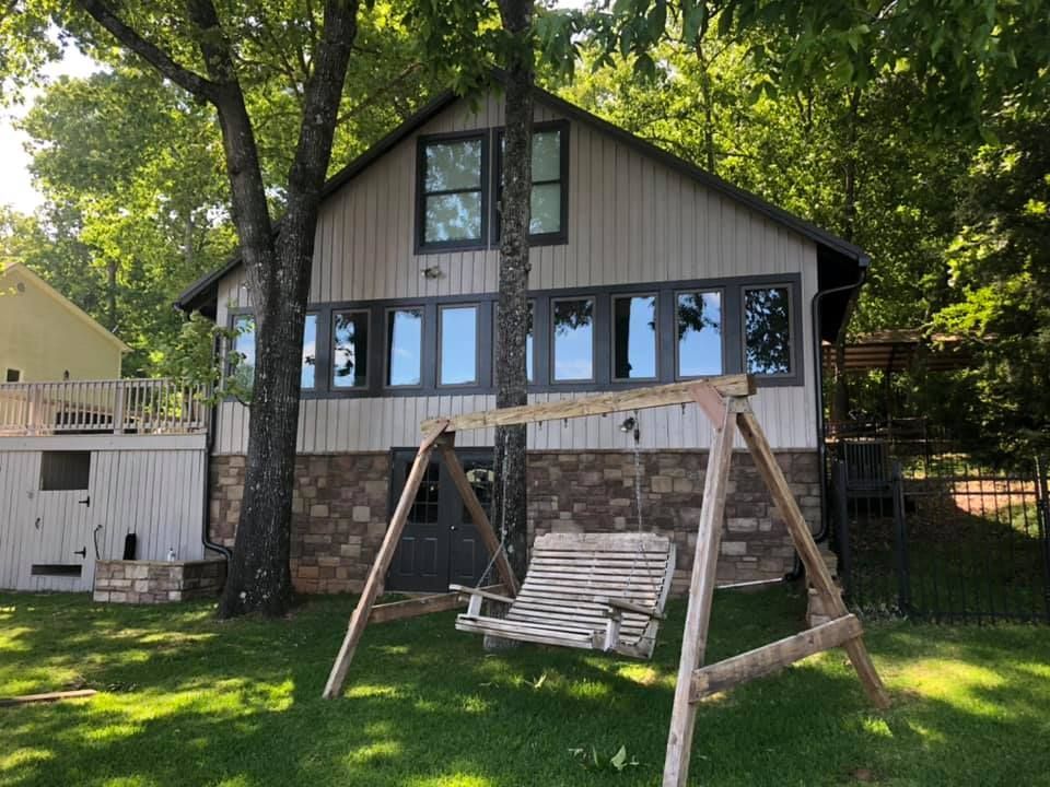 Cabin with stone foundation and tan siding, swing set in front, tall trees and green grass.