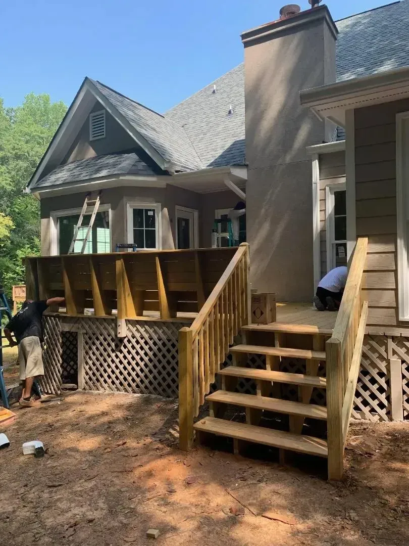 Construction of a wooden deck with stairs attached to a house; workers are present, outdoors.
