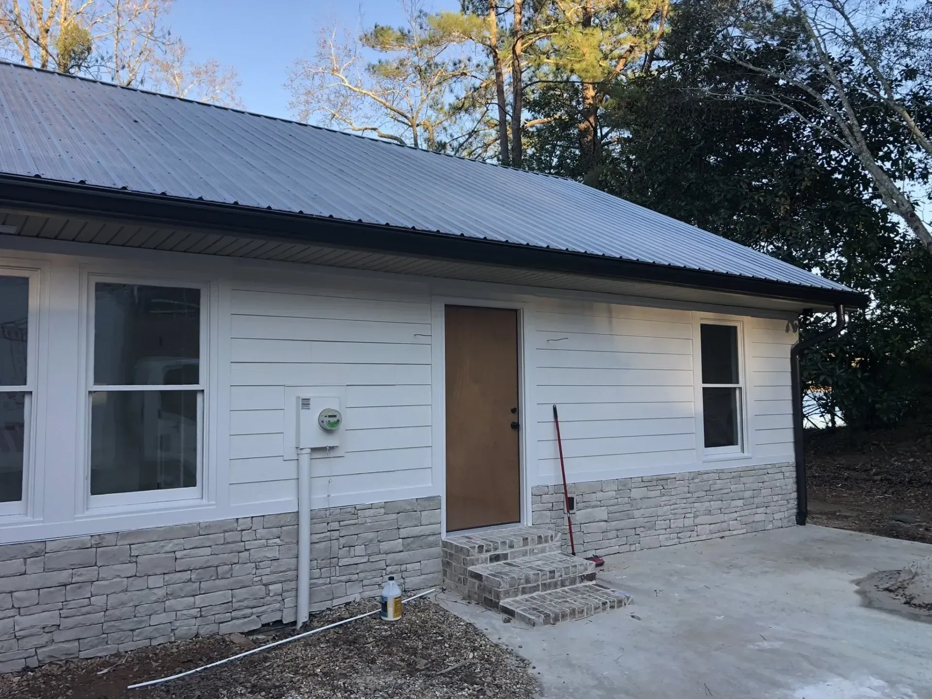 White-sided building with gray metal roof, brick base, brown door, and two windows.