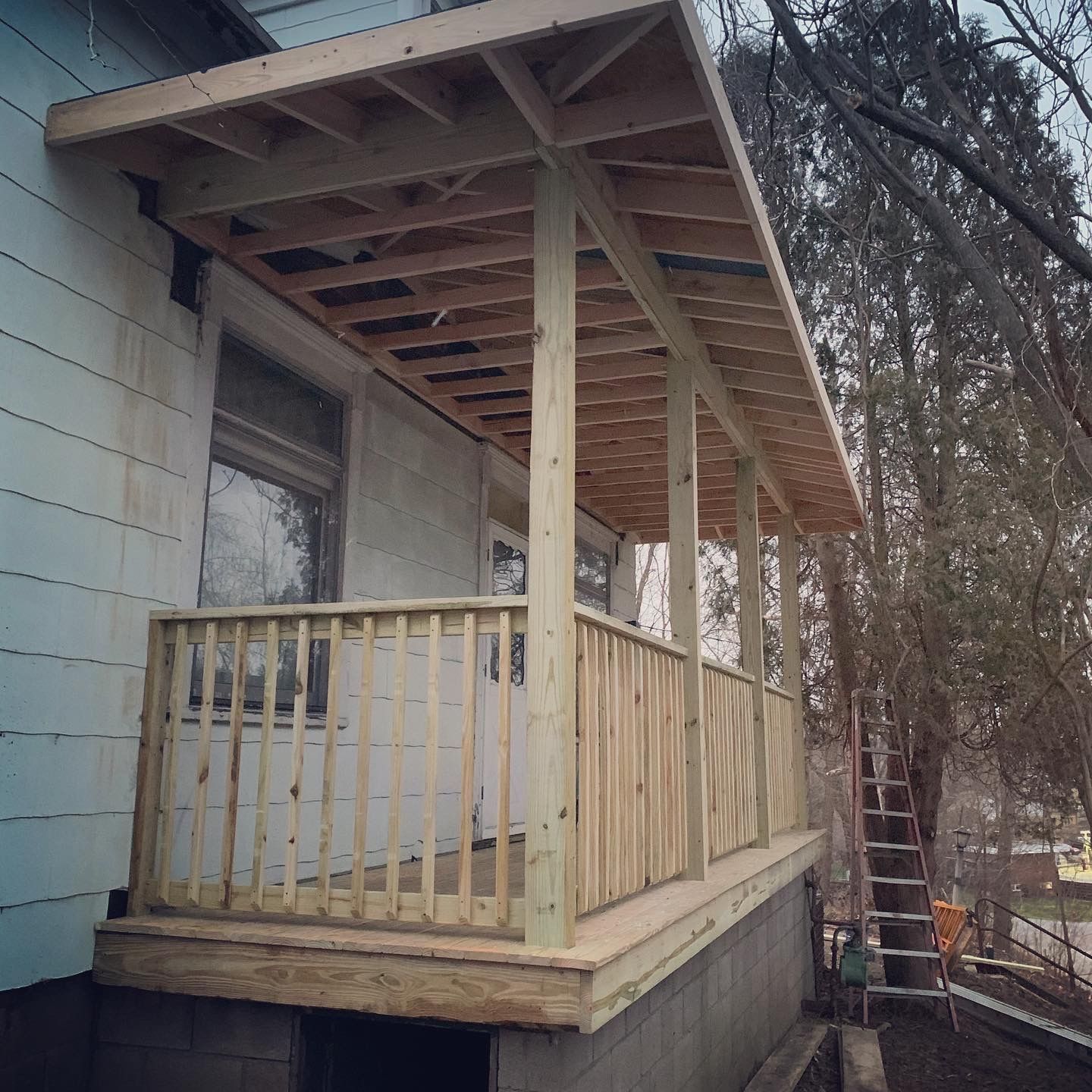 New wooden porch under construction with railing and roof attached to a white house.