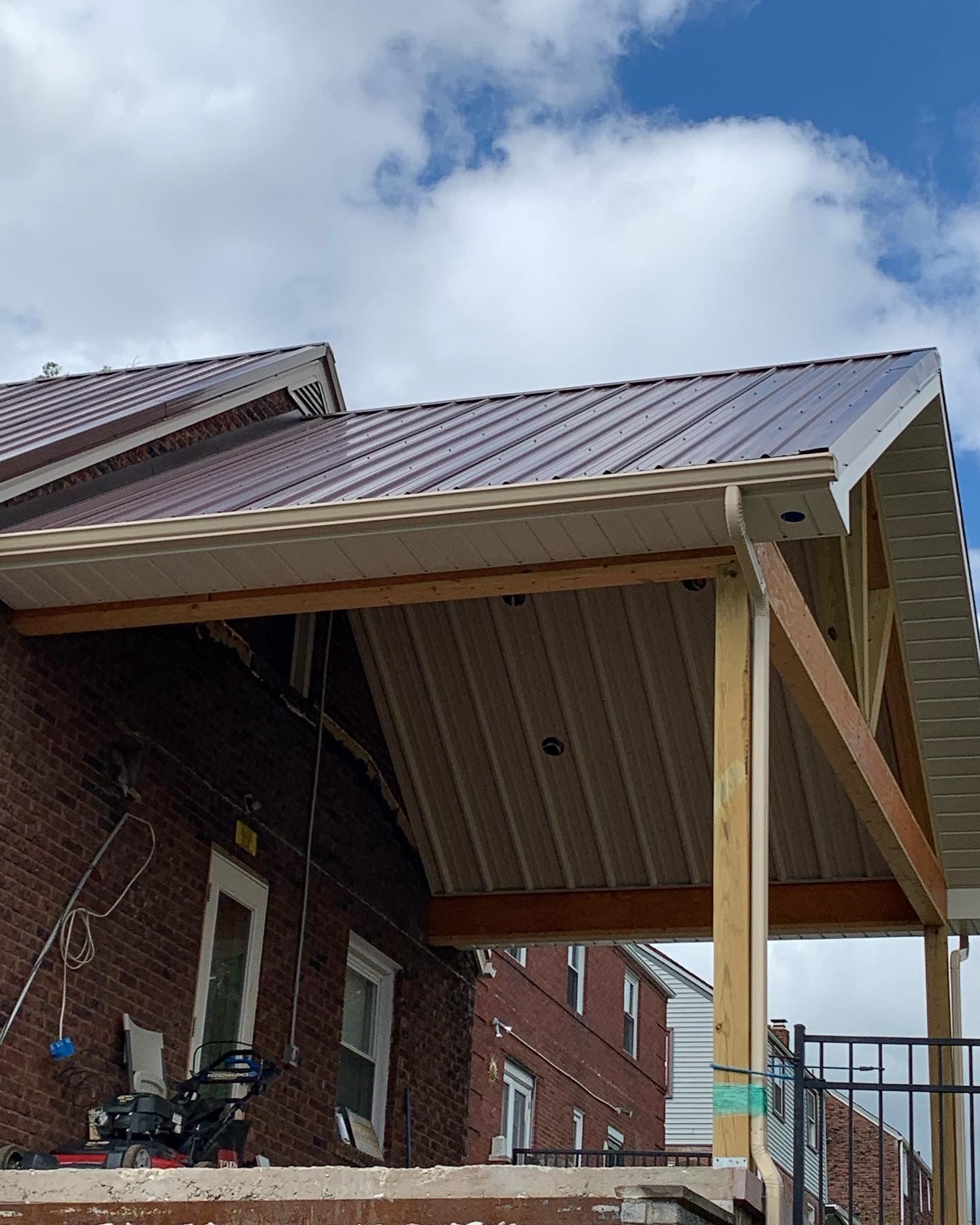 Red brick building with a brown metal roof, partially covered by a beige-colored porch. Blue sky with clouds.