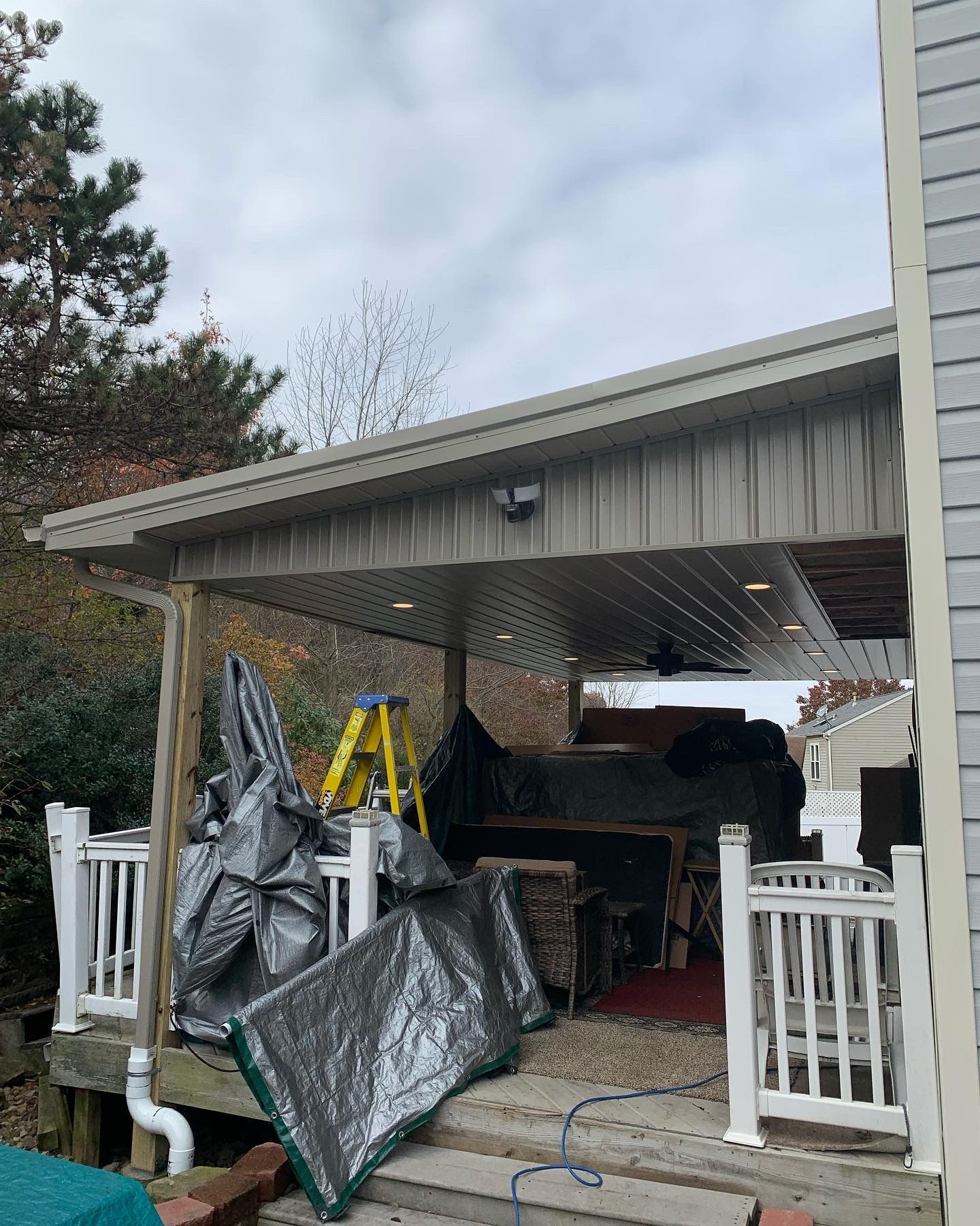 Covered deck with white railings, siding, and a covered area with tarps and construction materials under a cloudy sky.