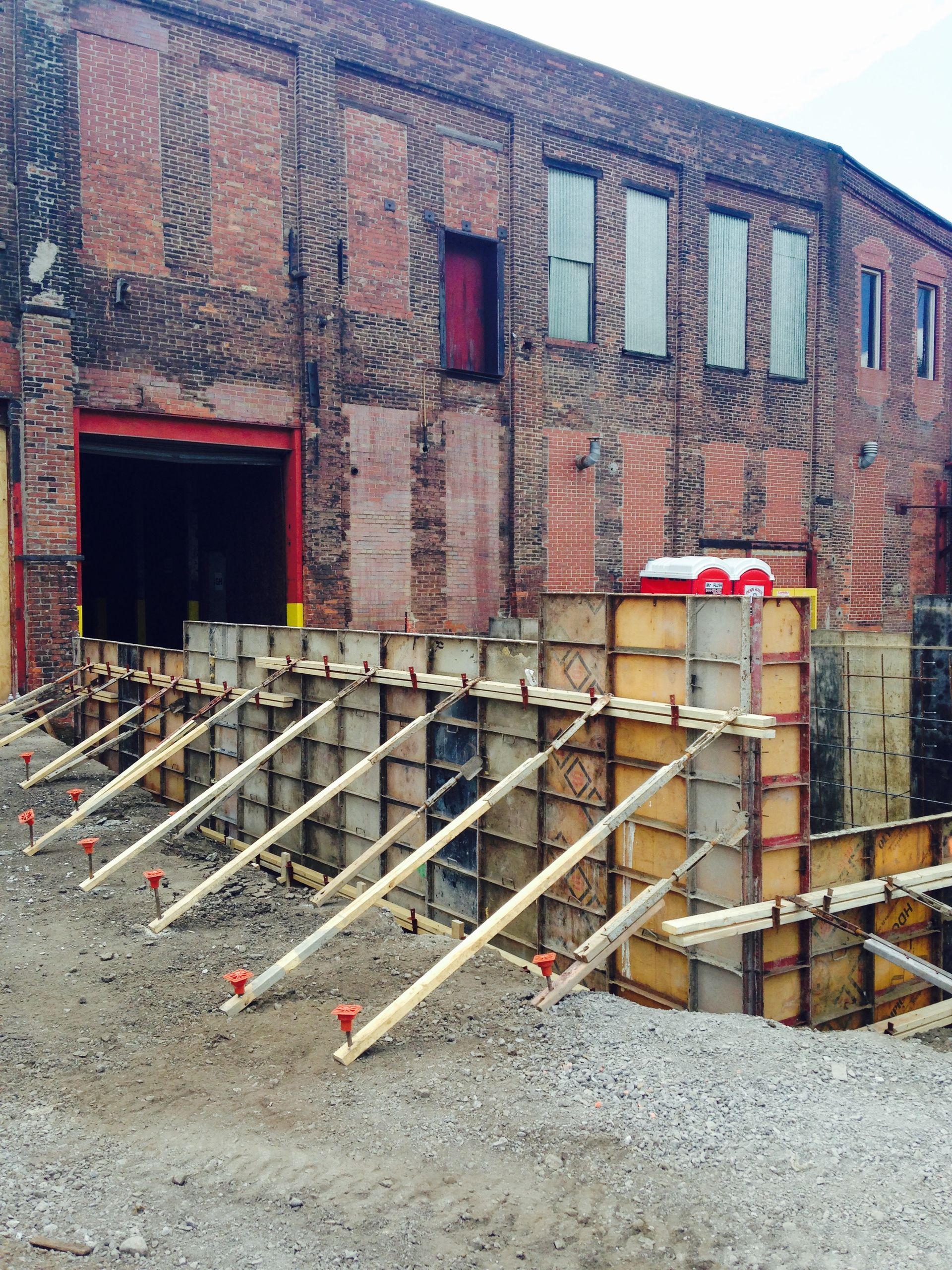Construction site with wooden forms supported by angled braces in front of a brick building.