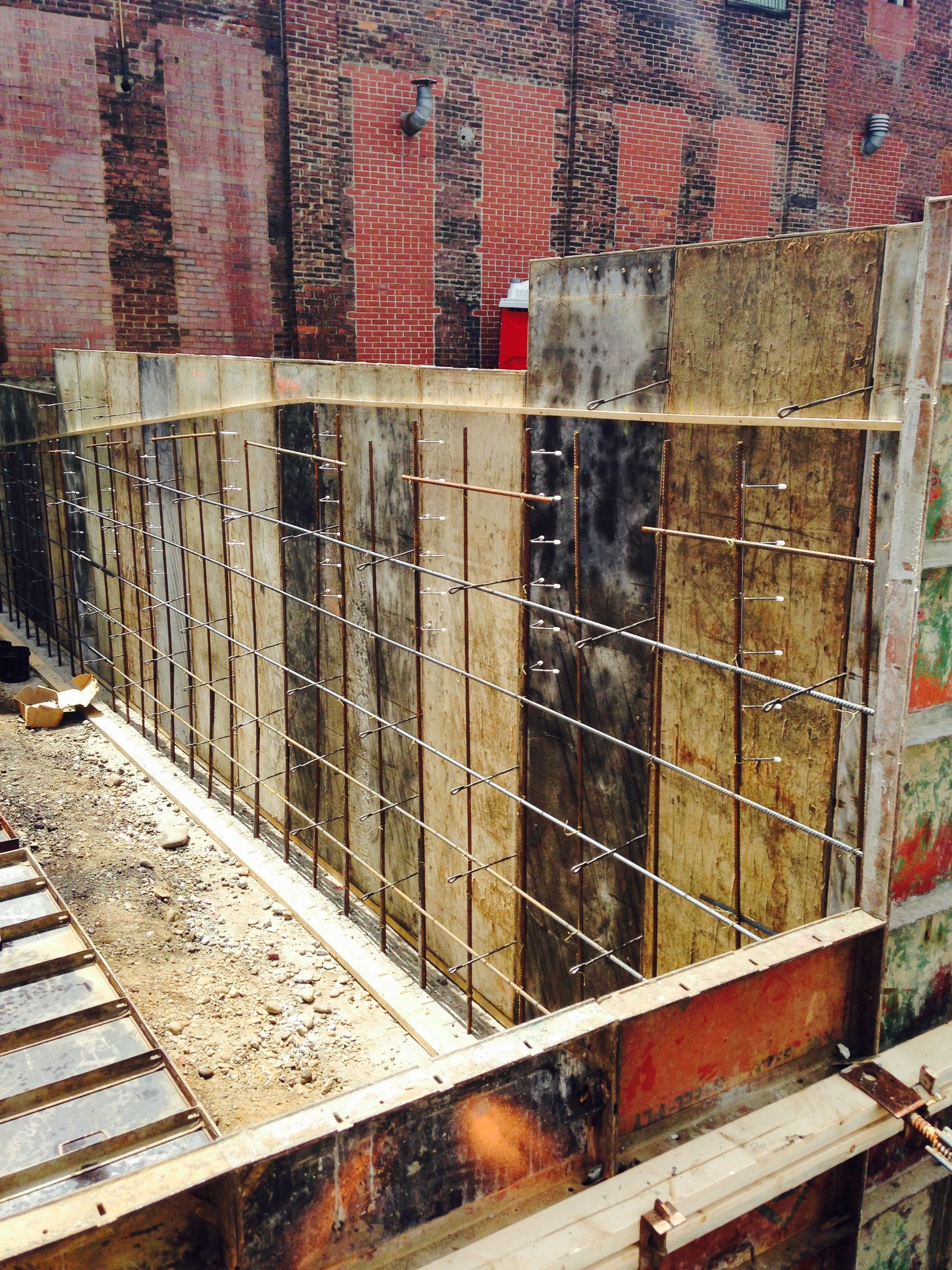 Construction site with exposed rebar, wooden forms, and a brick wall.