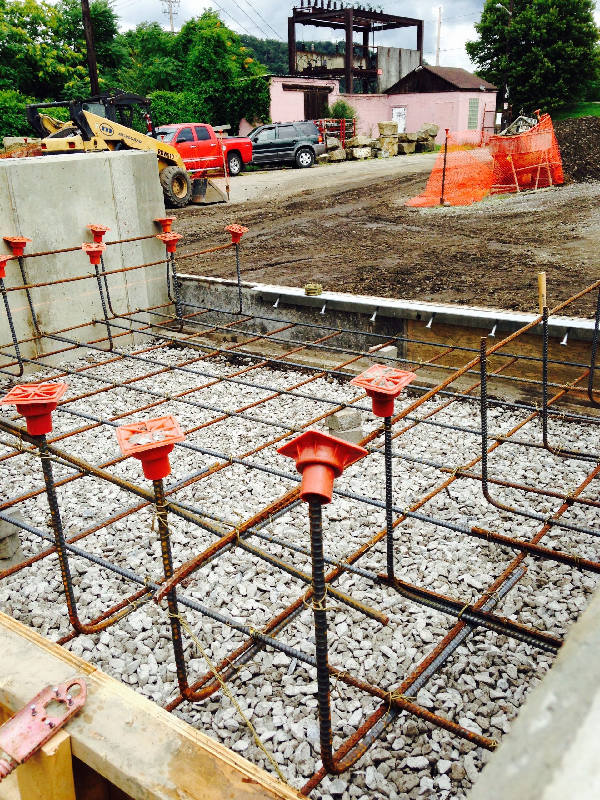 Construction site: Rebar grid with orange caps, gravel base, wooden frame, red truck in background.