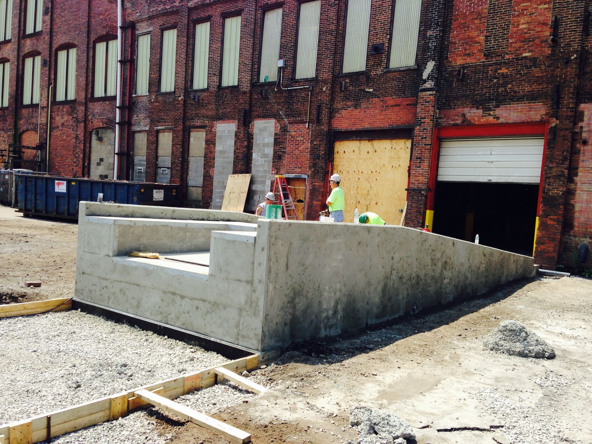 Concrete construction of a ramp and retaining wall outside a brick building. Construction workers are present.